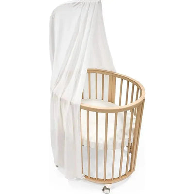 White canopy draped over a circular Stokke Sleepi crib with wooden slats and a soft mattress inside.