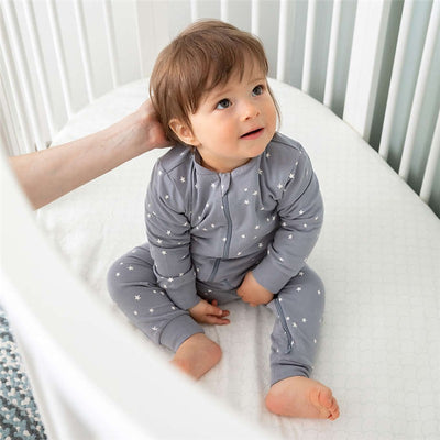 A toddler in a gray star-patterned onesie sits in a crib with a fitted sheet, smiling as a hand reaches toward them.