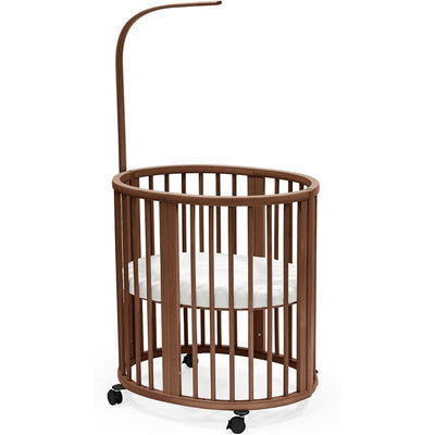 Stokke® Sleepi™ Mini Bundle in Warm Brown, featuring a round wooden crib with a removable white mattress and wheels for mobility.