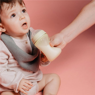 Light blue Suavinex Color Essence SX Pro™ silicone bottle held by an adult, with a surprised baby sitting and wearing a gray sweater.