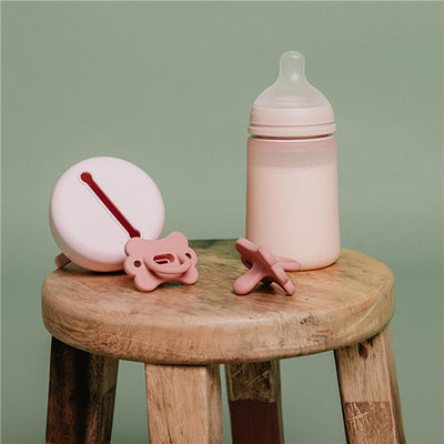 Suavinex Color Essence SX Pro™ Silicone Bottle 8 oz in rose water next to a pink pacifier and a round container on a wooden stool.