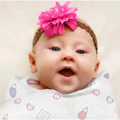 Baby smiling, wrapped in a Swaddle Designs Marquisette Swaddle Blanket with a peace and love print, wearing a pink flower headband.