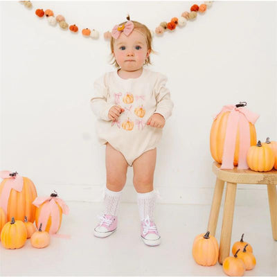 Baby girl wearing a pumpkin-themed outfit and pink headband, surrounded by decorative pumpkins and a wooden stool.