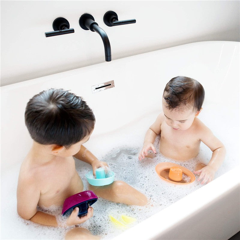 Two children playing in a bubble bath, each holding colorful stacking boats; one is navy blue and the other light blue.
