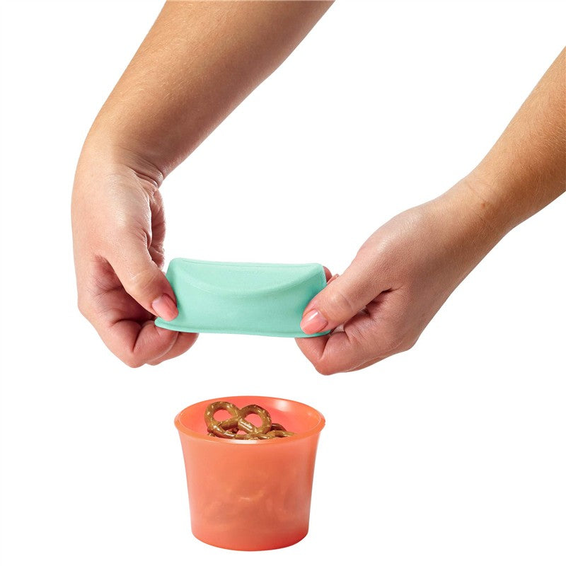 Hands holding a mint-colored silicone lid above a clear orange container filled with pretzels.
