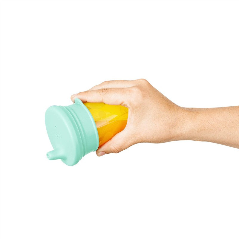 Boon SNUG Spout Universal Silicone Sippy Lid in green, being held over a yellow cup, designed for spill-proof sipping.