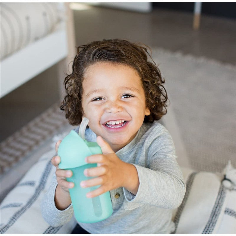 Child smiling while holding a mint green Boon SWIG Silicone Straw Cup, 9 oz, with a straw, in a cozy indoor setting.