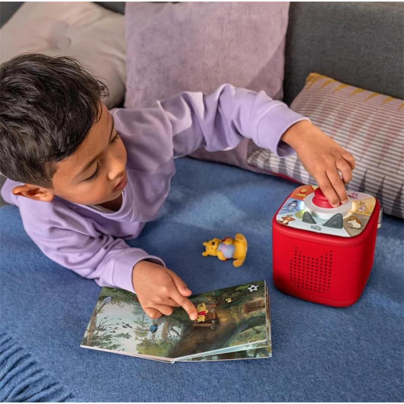 Young child interacts with a red Toniebox and points at a storybook while a Winnie the Pooh figure is beside them.
