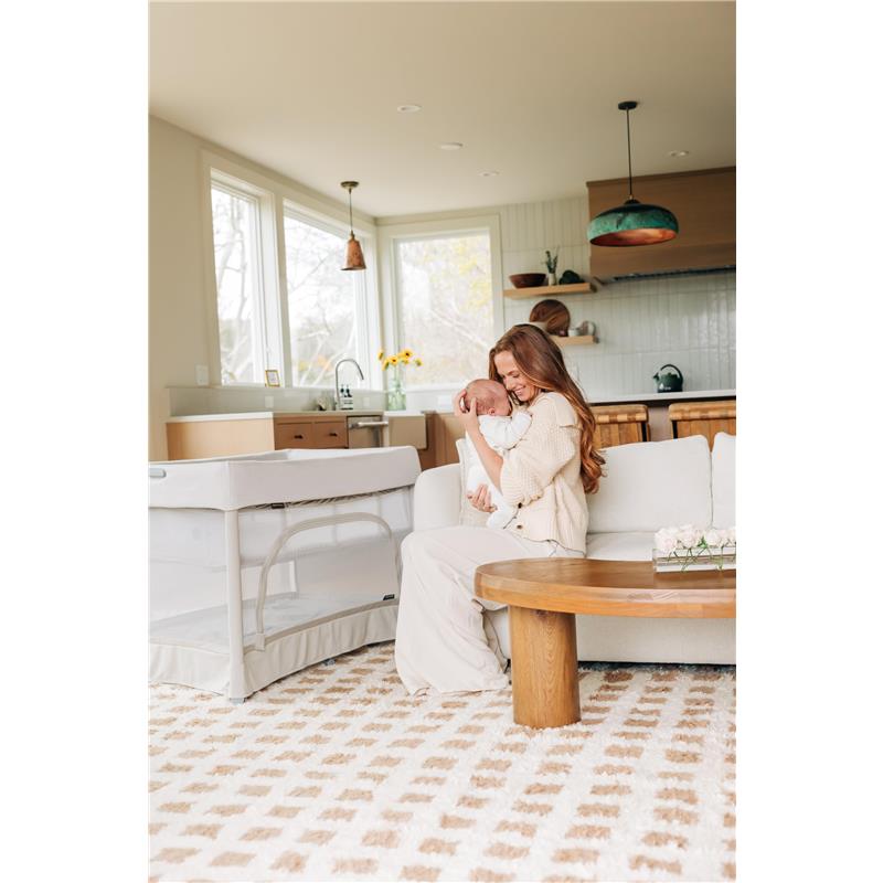 Mother holding a newborn in a cozy living room with a UPPAbaby Remi V2 Playard nearby.