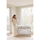 Mother holding a baby in a cozy room next to a UPPAbaby Remi V2 Playard, featuring a waterproof mattress cover.