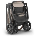Uppababy Kona stroller in Sandstone Mélange, featuring a compact folded design with a dark frame and light fabric accents.