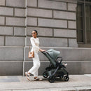 A woman walks with the Uppababy Kona stroller in green, wearing sunglasses and casual white attire, against a city backdrop.