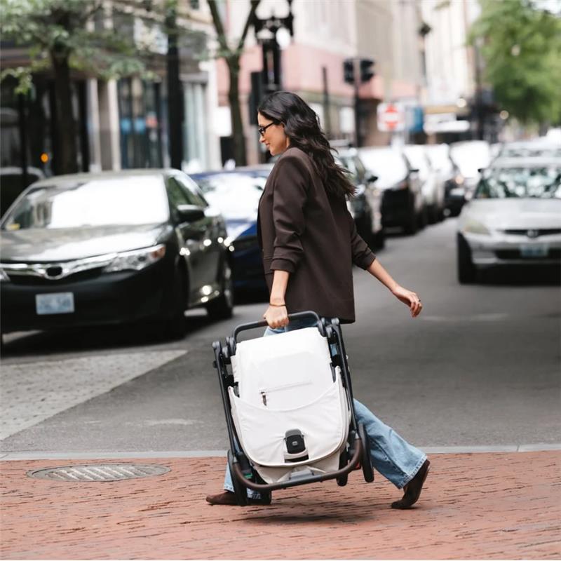 A woman walks down a city street carrying a folded Uppababy Kona stroller and bassinet in Rowan (Graphite) color.