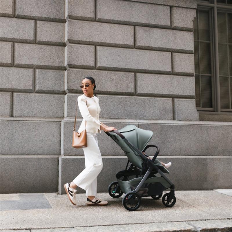 Woman in a stylish outfit walking with the Uppababy Kona stroller in James (Black) color against a stone wall.