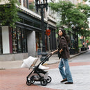 A woman pushes an Uppababy Kona stroller with a Mesa V3 car seat attached, strolling down a city street.