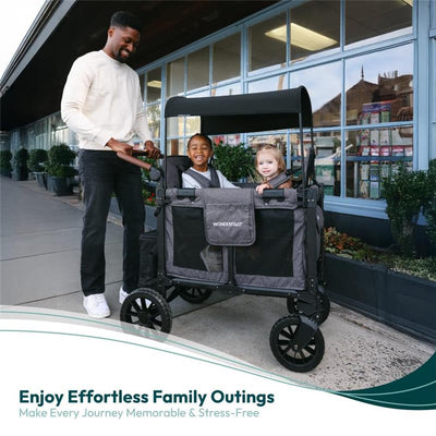 Two children smile from inside a Wonderfold W2 Luxe Pro Stroller Wagon while an adult pushes it along a sidewalk.