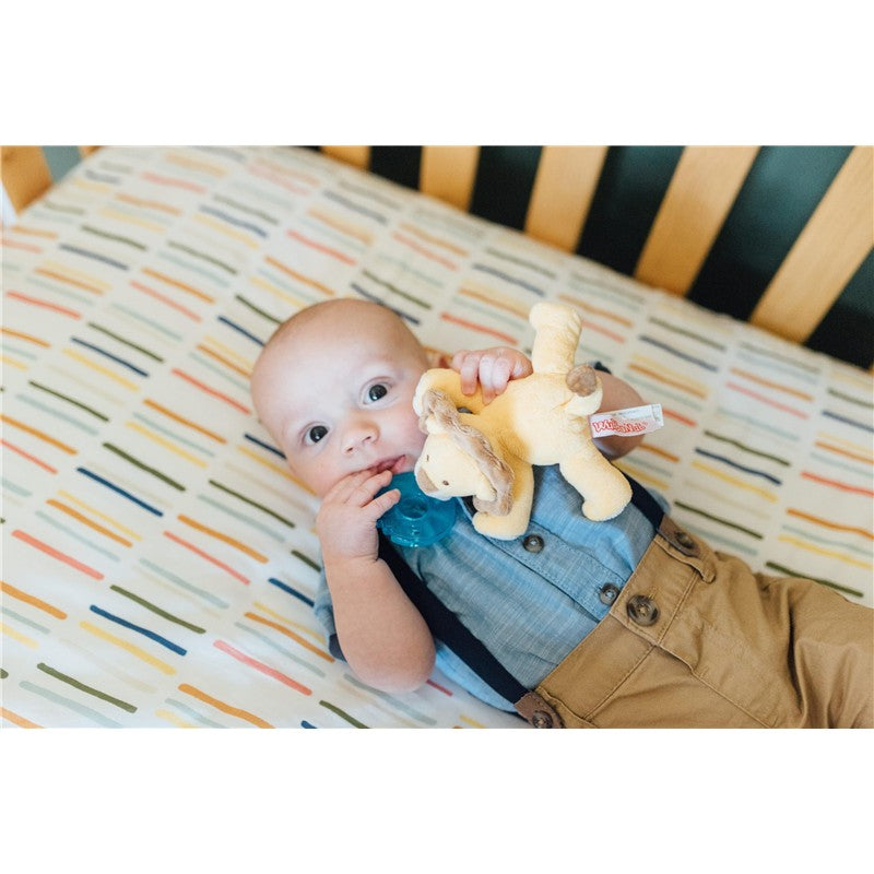 Infant holding a blue pacifier and a plush baby lion while lying on a crib sheet with colorful stripes.