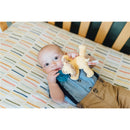 Infant holding a blue pacifier and a plush baby lion while lying on a crib sheet with colorful stripes.