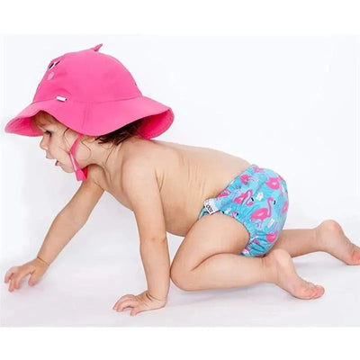 Toddler crawling, wearing a pink sunhat and a blue swim diaper with flamingo patterns, against a white background.